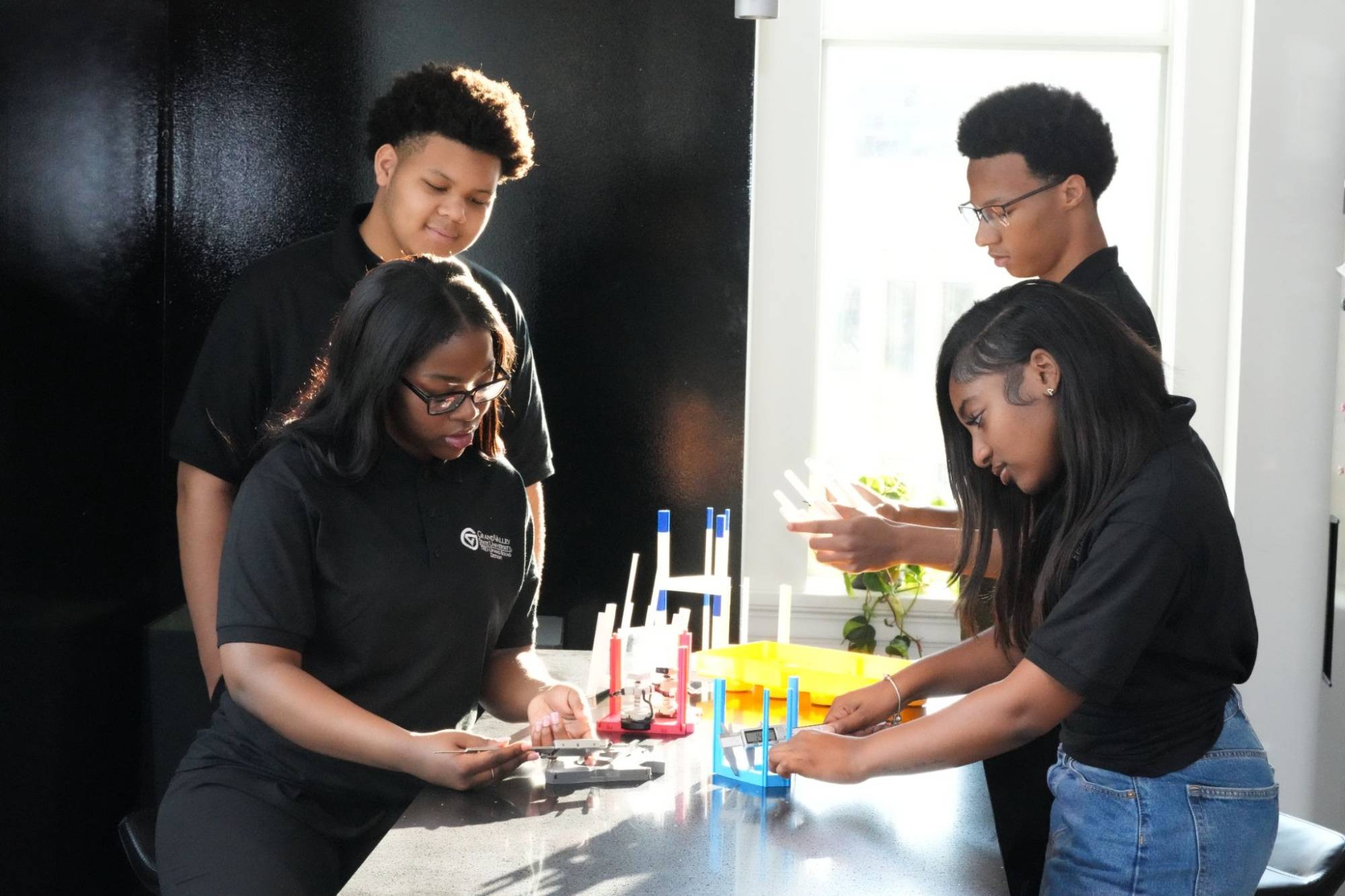 Four students wearing matching black shirts work together on a hands-on project at a lab table.